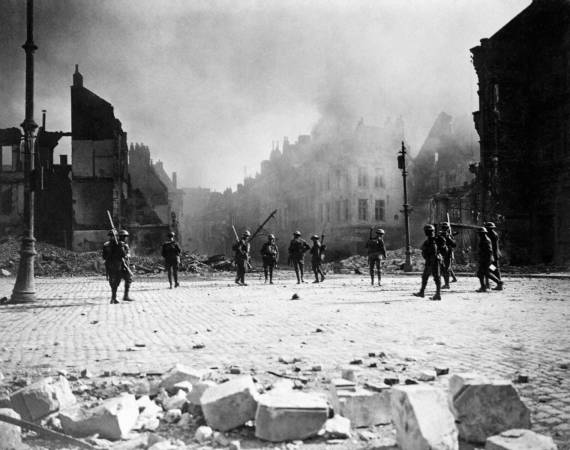A group of Canadian soldiers strung out in the streets of Cambrai against its still burning buildings